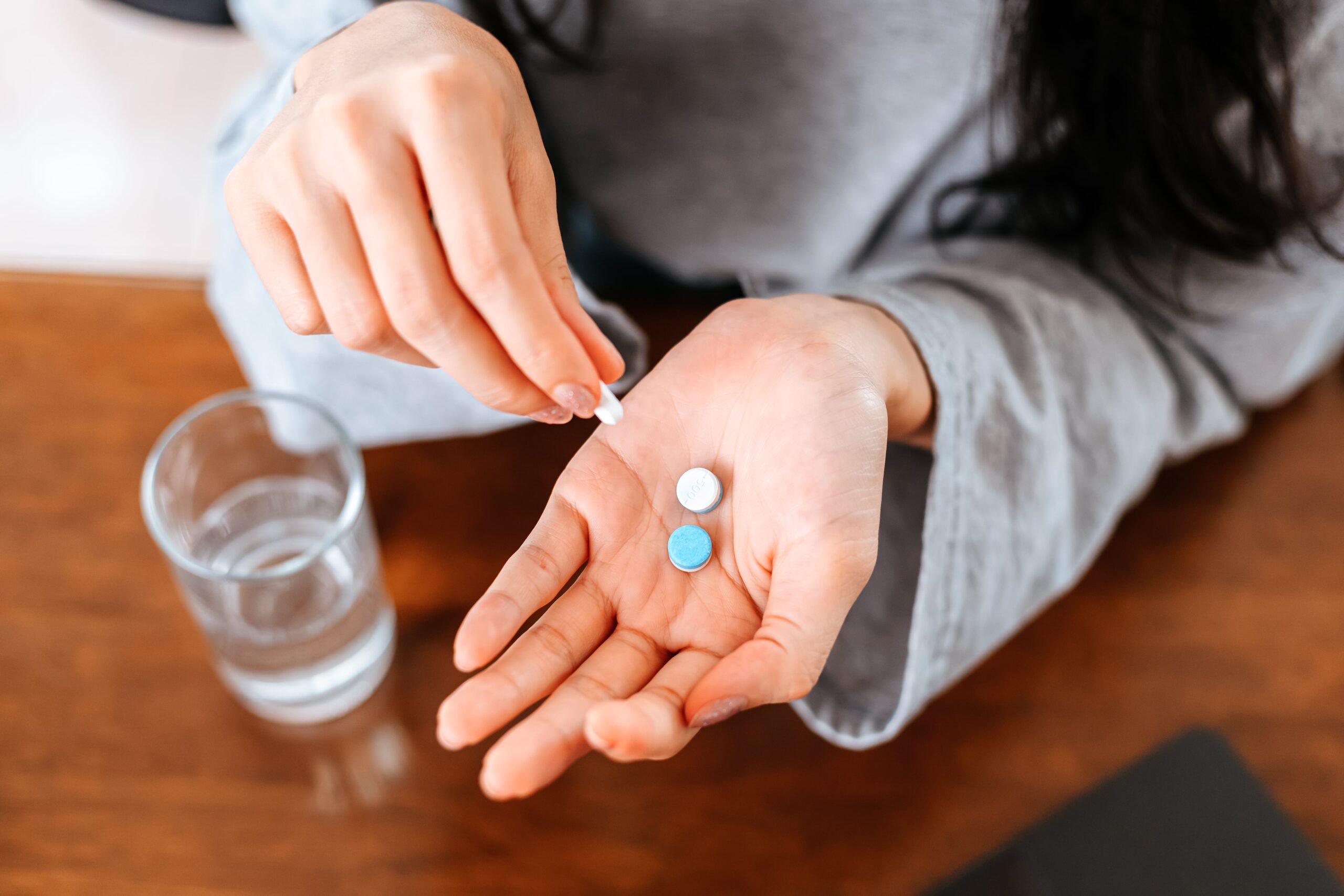 A person holding pills with a glass of water on the table.